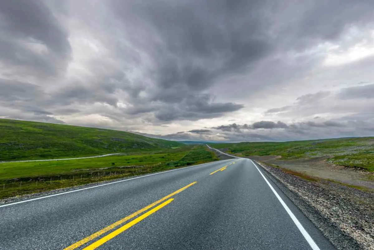 Straight road through rolling green hills and rocky tundra, under heavy gray clouds.