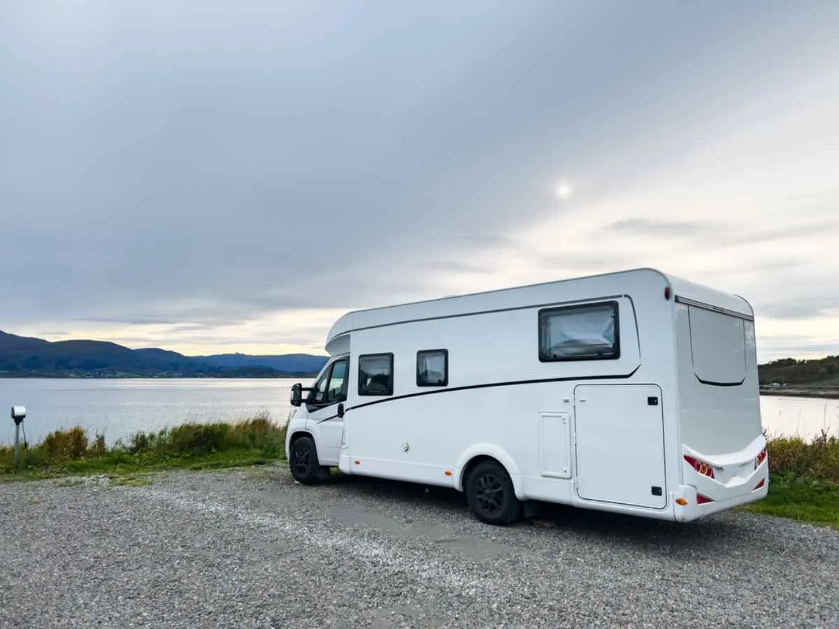 White motorhome parked at a waterfront campsite with calm water and distant hills near Ålesund, Norway.