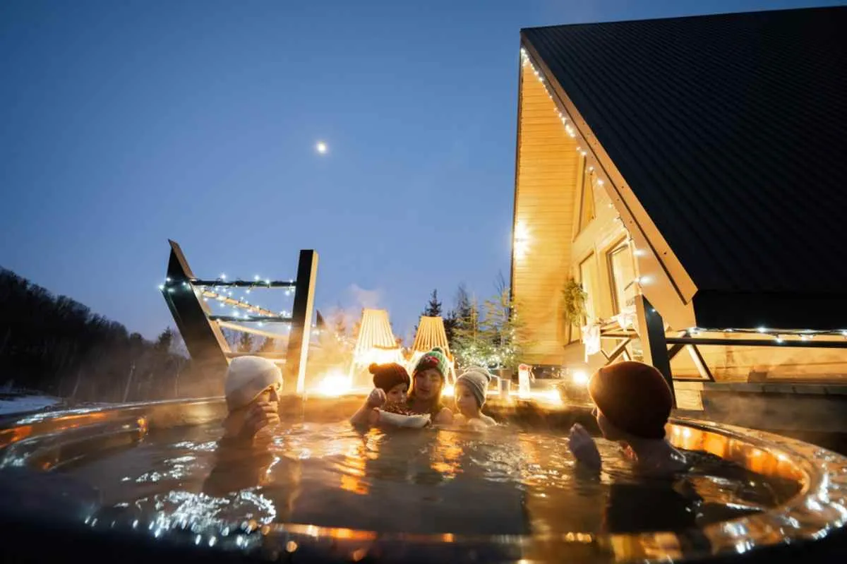 Family relaxing in a steaming outdoor hot tub beside a cabin strung with lights at dusk.