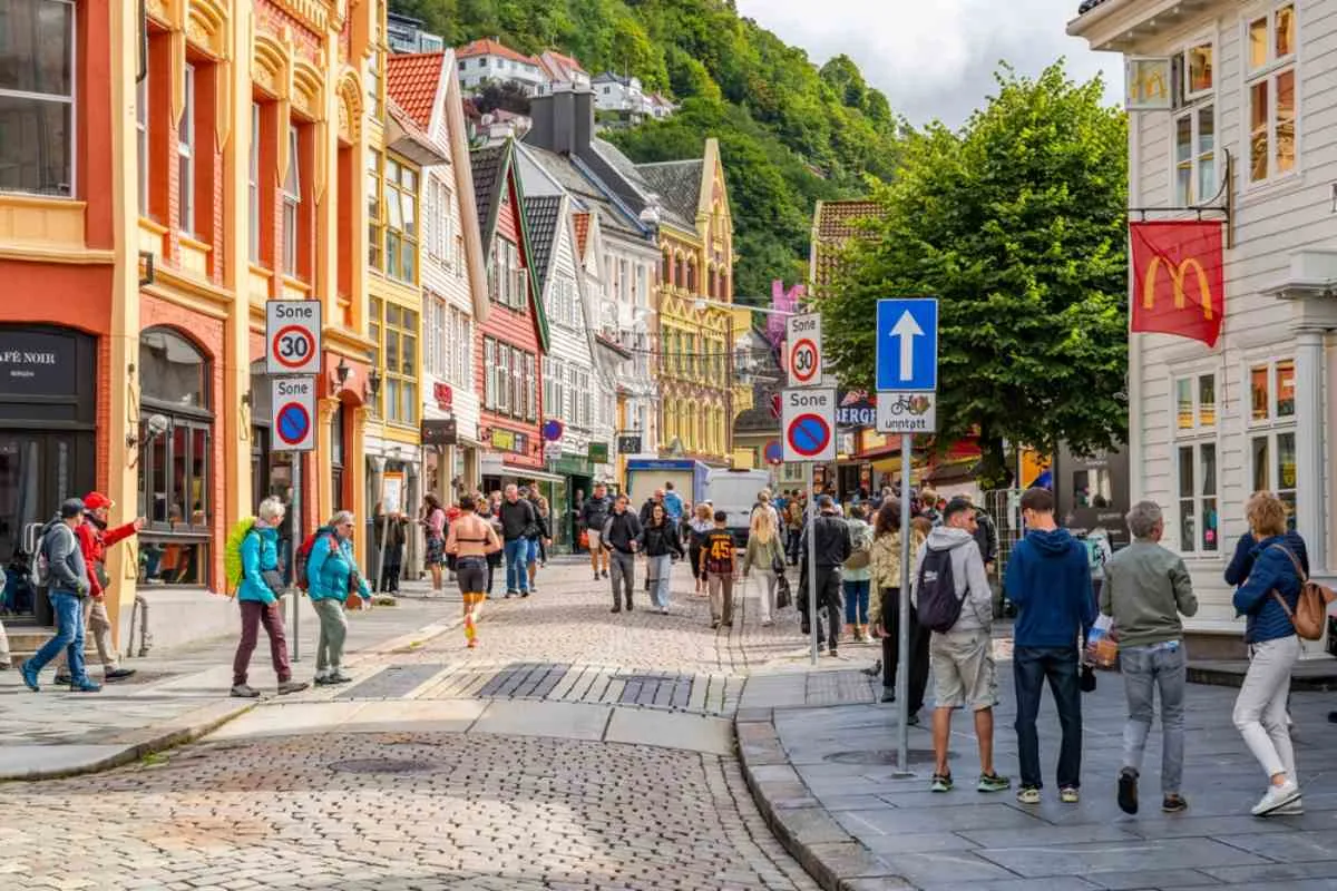 Colorful wooden houses and a cobbled street in Bergen’s historic center, pedestrians strolling beneath speed signs near Bryggen on a bright day.