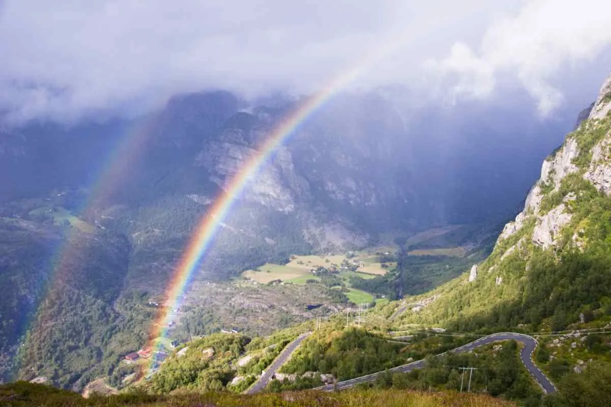 Double rainbow arches over a Norwegian mountain valley, winding road and farms below, misty cliffs and sunlit clouds after a passing storm.