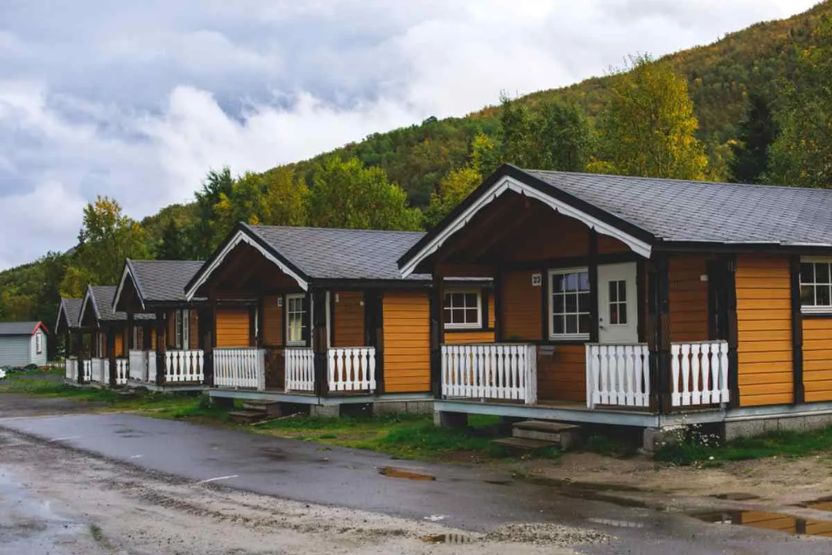 Row of cozy wooden camping cabins with white railings in Norway, puddled driveway and forested hillside behind under overcast autumn skies.