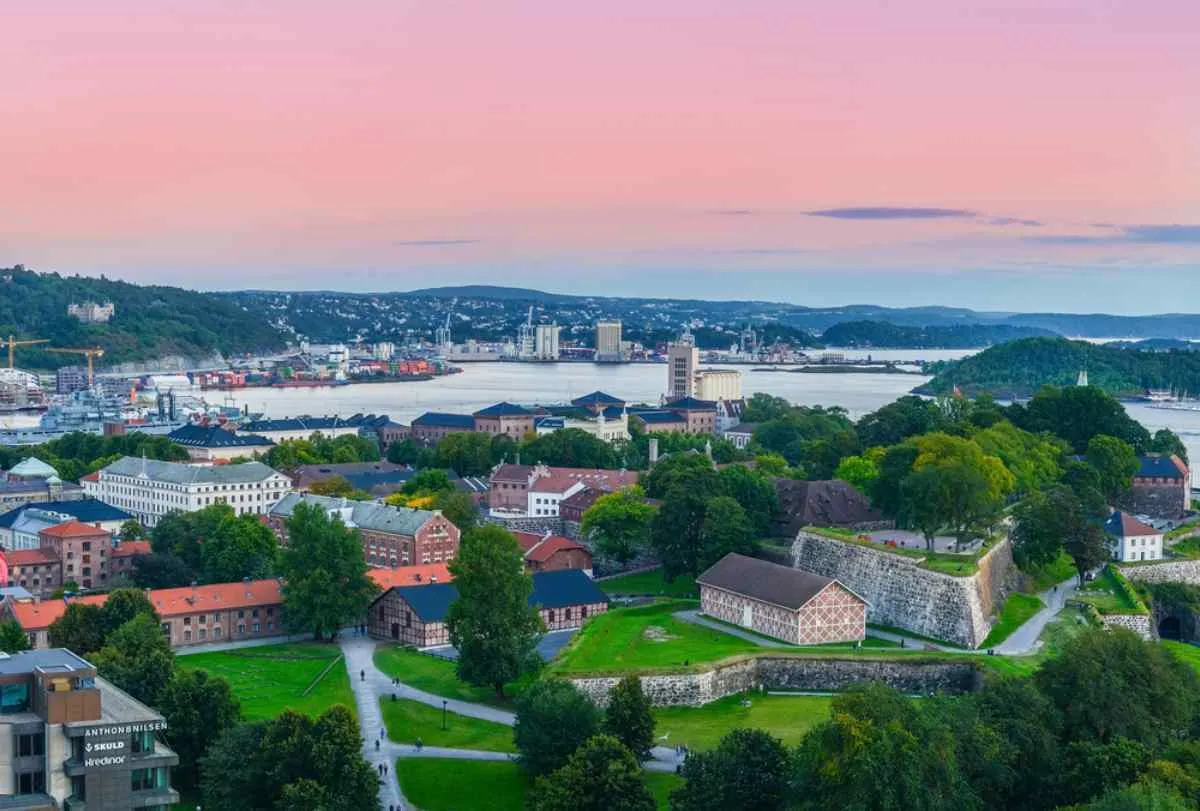 Panoramic view of Oslo and the harbor at sunset, Akershus Fortress and green parks in the foreground, islands and city skyline around the bay.