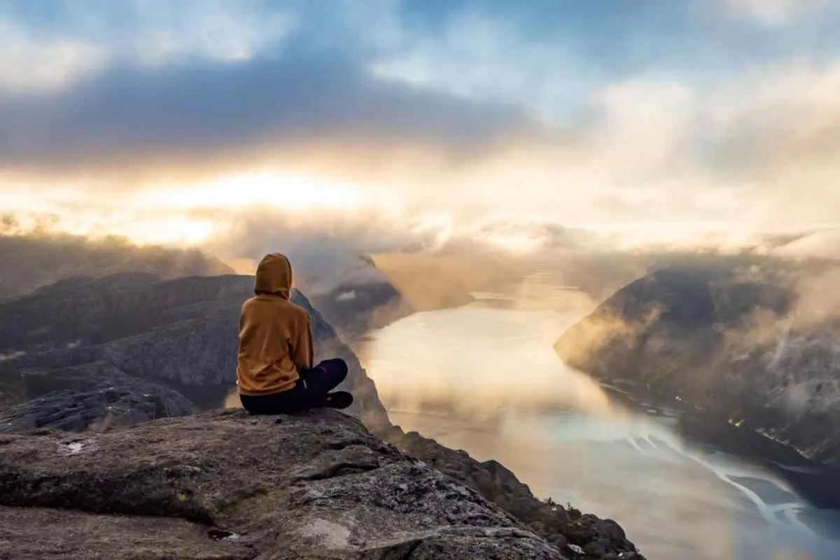 Hiker in a hooded jacket sits on the edge at Preikestolen, watching sunrise light spill over Lysefjord’s cliffs and winding water below.