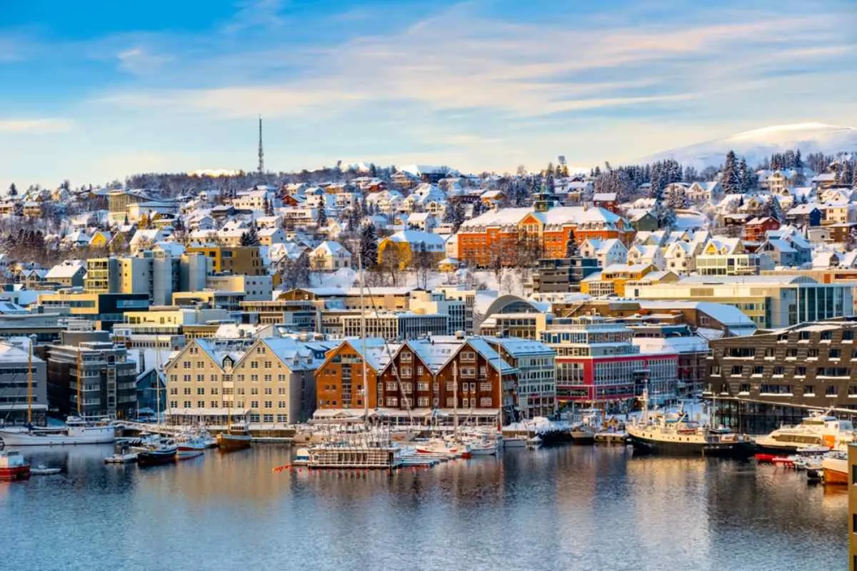 Winter view of Tromsø harbor with snow-covered rooftops, wooden warehouses, and moored boats, soft morning light on the Arctic city and hills.