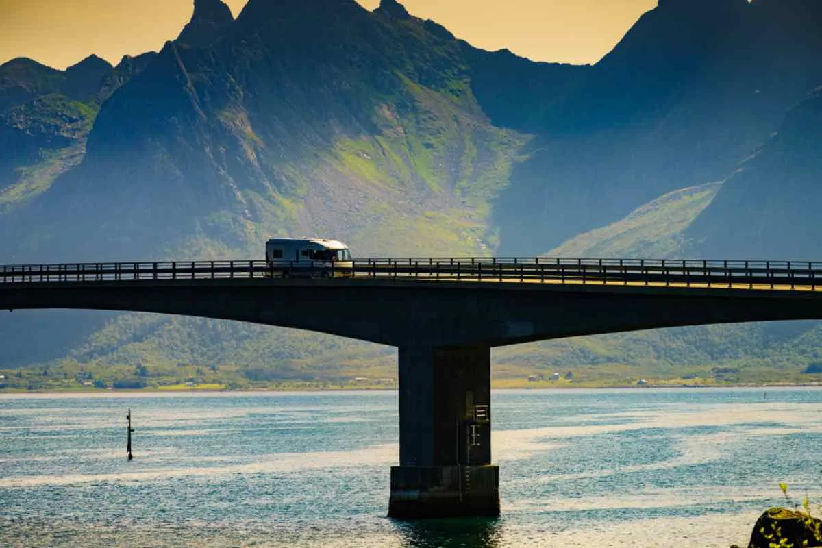 Campervan driving across a bridge in the Lofoten Islands, with fjords and rugged mountains in the background.