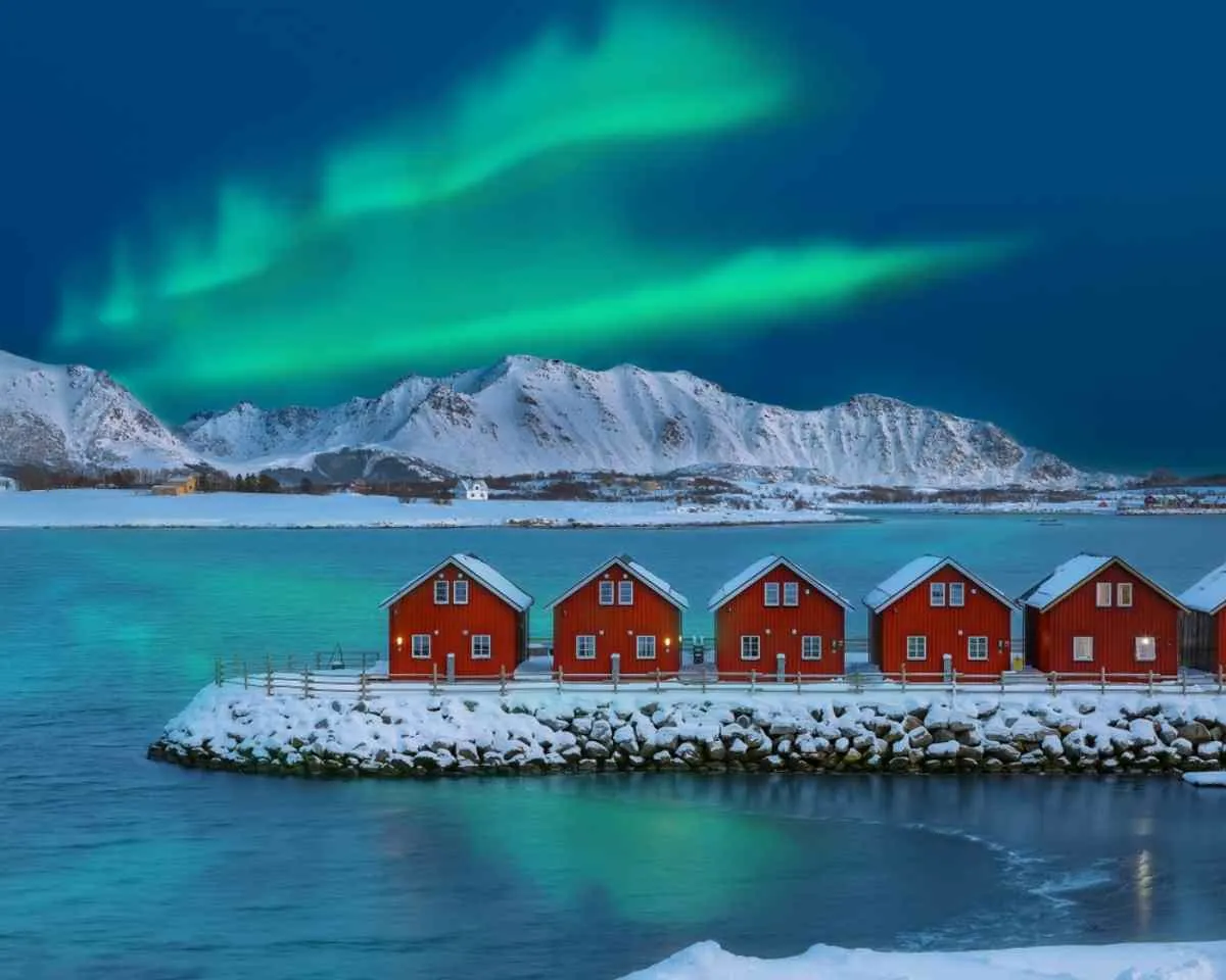 Northern Lights dancing above red fishermen’s cabins on the coast of the Lofoten Islands during winter.