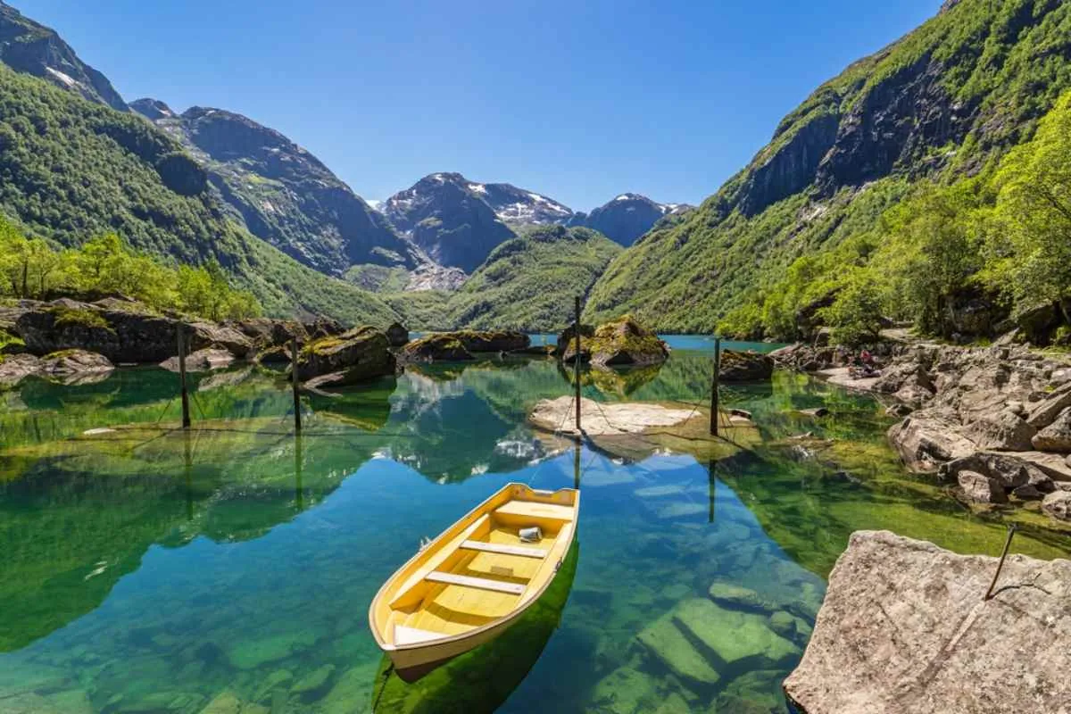 Yellow rowboat floating on crystal clear green water in a mountain lake, surrounded by rocky shores and steep forested peaks under a bright blue sky.