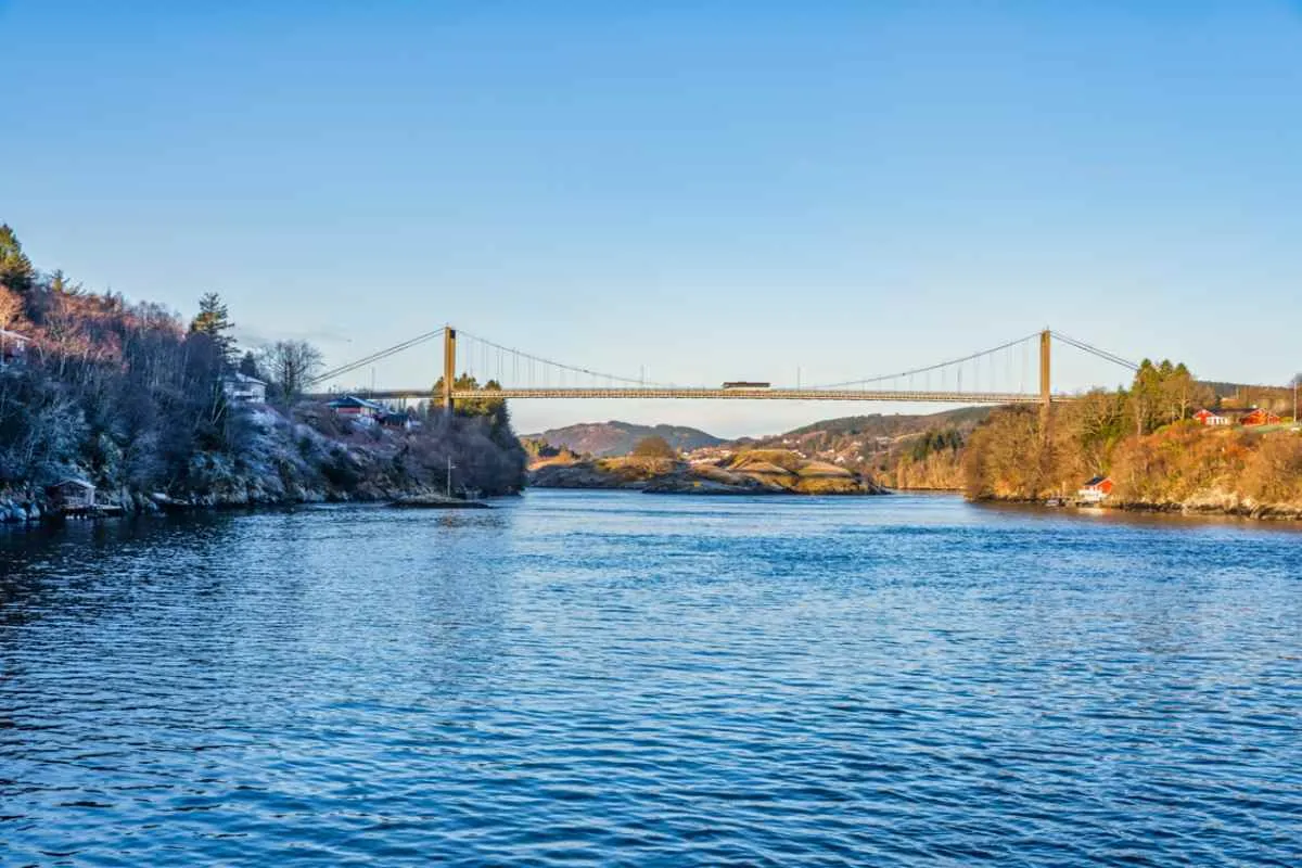 Suspension bridge crossing a calm fjord between rocky shores, with winter light, blue water, and scattered houses along the coastline.