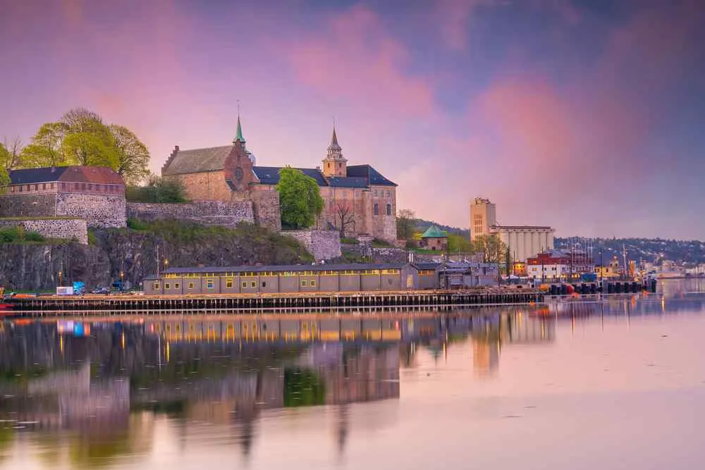 Akershus Fortress and Oslo waterfront reflected in calm water at sunset.