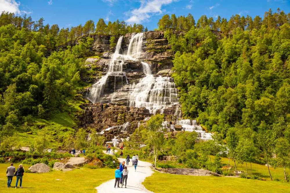 Visitors walking toward Tvindefossen waterfall surrounded by green forest in Norway.