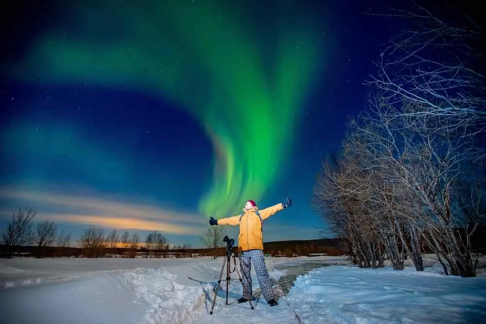 Traveler standing beside a camera tripod under vivid green northern lights over a snowy landscape in northern Norway.