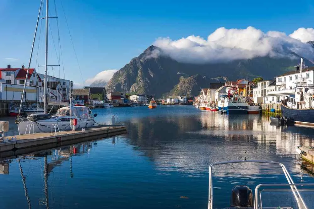 Fishing boats and colorful waterfront buildings in a peaceful Lofoten harbor surrounded by steep mountains and low clouds.