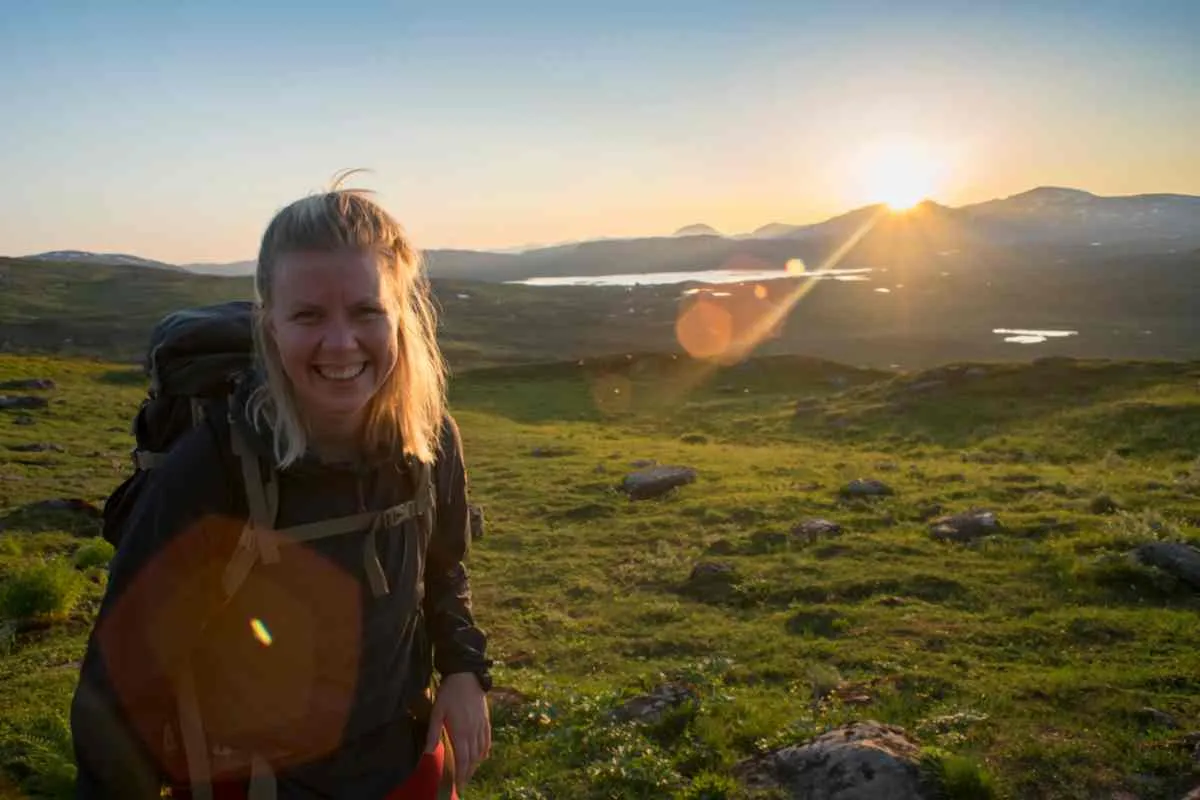 Une femme souriante en tenue de randonnée, un sac à dos sur le dos, avec le soleil derrière elle.
