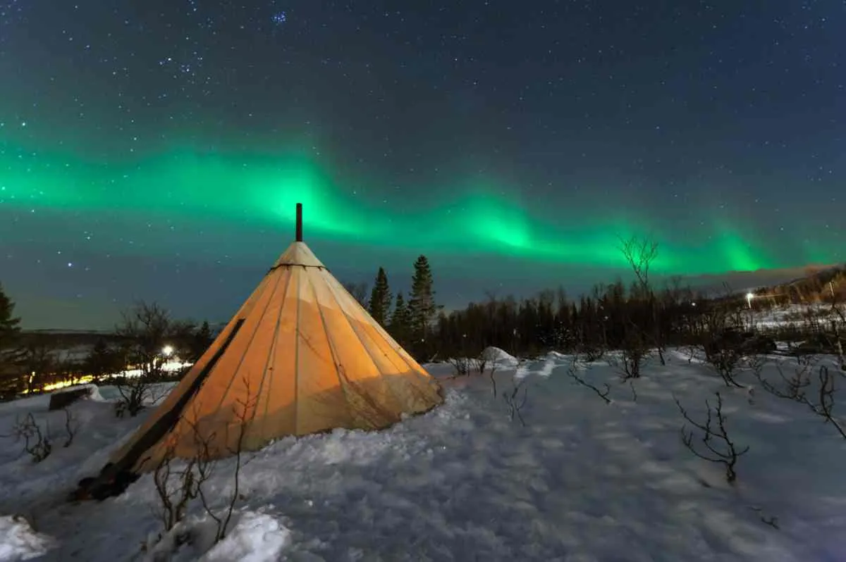 Une tente sami baignée d’une lumière chaleureuse dans une forêt enneigée, sous des aurores boréales d’un vert éclatant et un ciel étoilé.