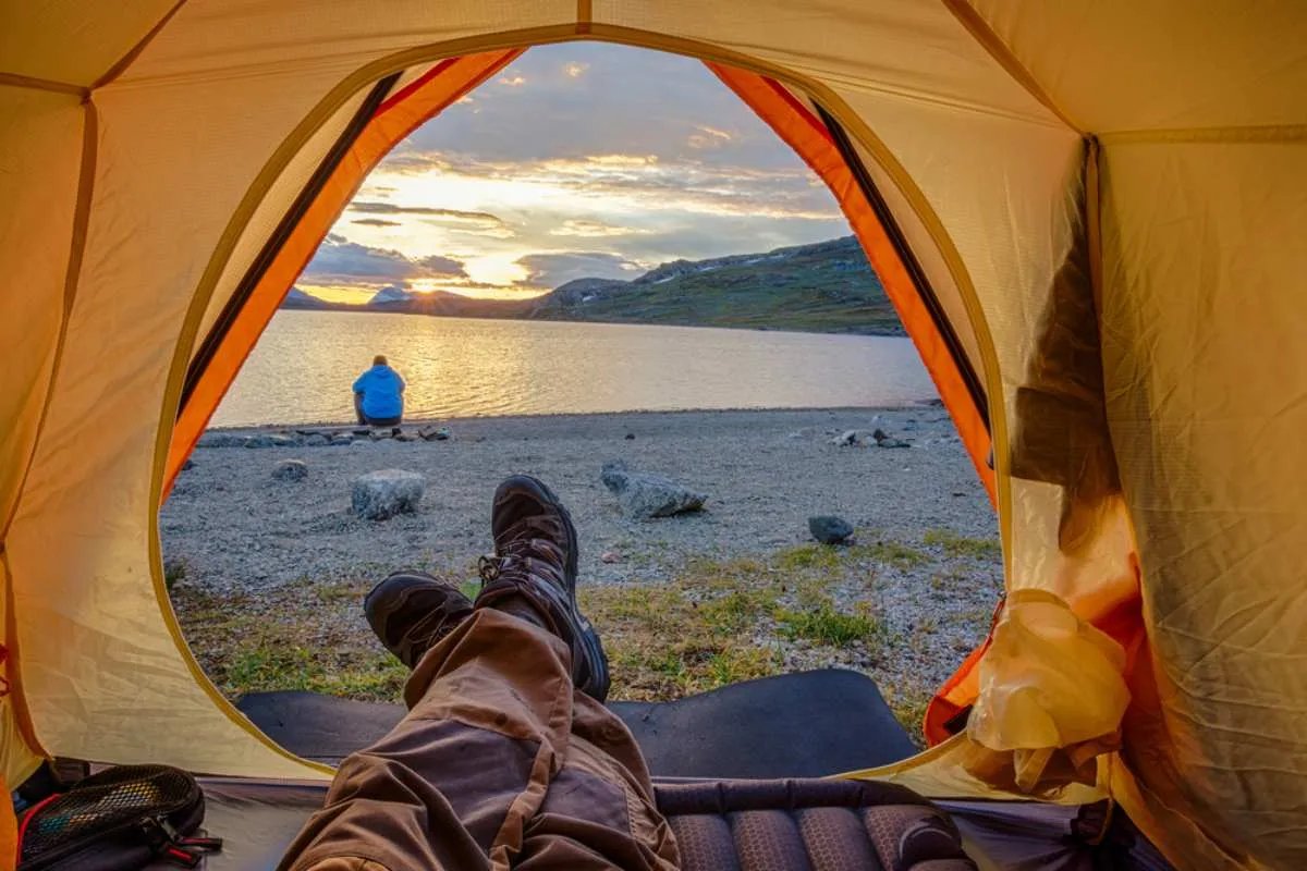 Vista desde el interior de una tienda de campaña hacia la tranquila orilla de un lago al atardecer, con unas botas de montaña en primer plano y una autocaravana aparcada junto al agua.