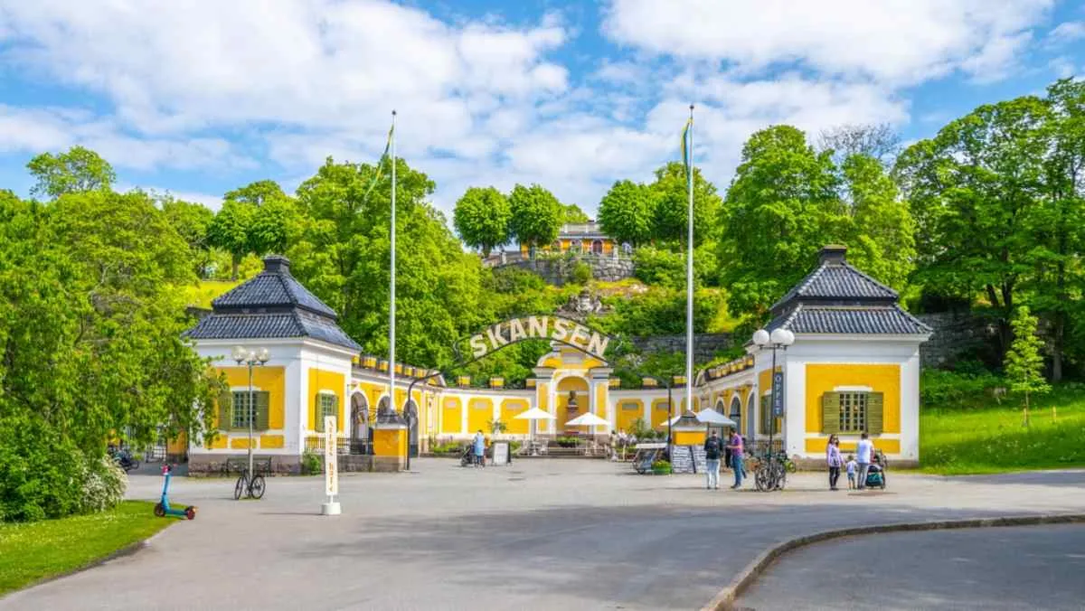 Vista general de la entrada principal de Skansen en Estocolmo, Suecia, con una fachada especialmente amarilla y vegetación verde a su alrededor.