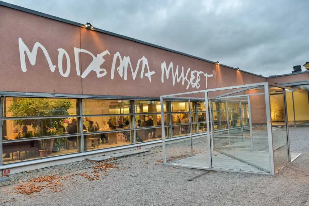 Vista general de la fachada del Moderna Museet de Estocolmo, con varias personas sentadas en lo que parece ser un comedor iluminado por la cálida luz que entra desde el interior a través de grandes ventanales.
