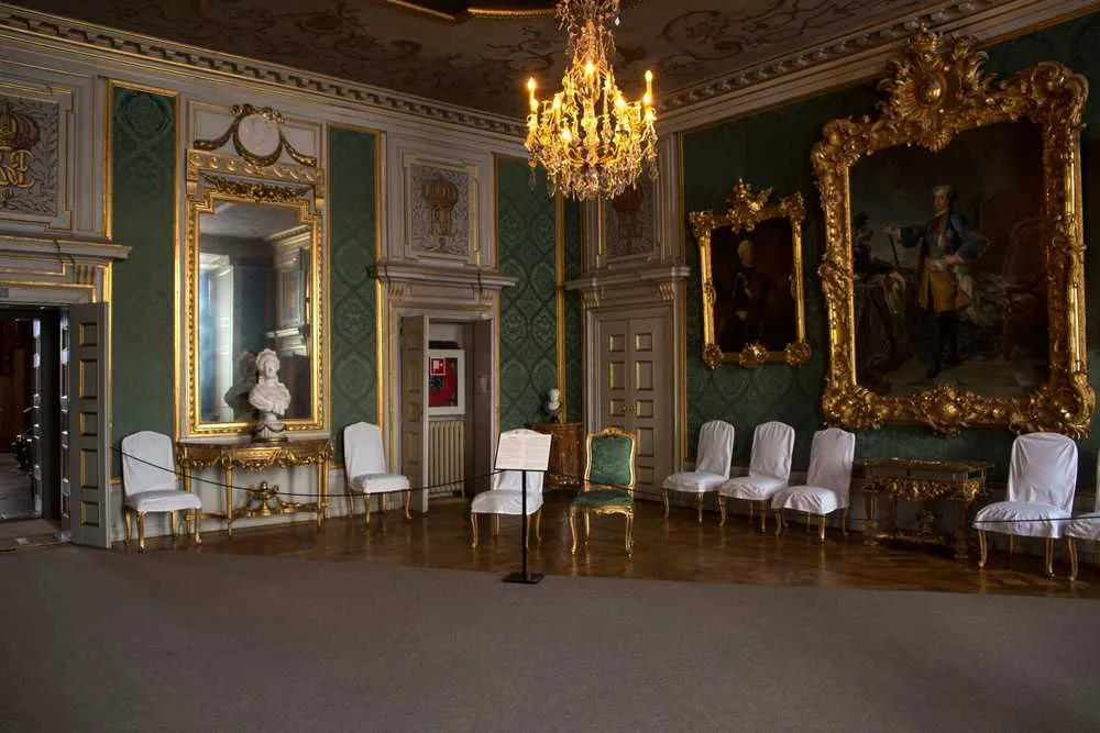Vista detallada de una de las salas del Palacio Real de Estocolmo, con varias sillas tapizadas, cuadros enormes con marcos dorados y un espejo delante del cual se encuentra una escultura blanca.