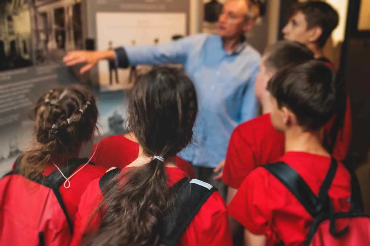 Primer plano de un niño vestido de rojo que escucha con atención las instrucciones de un señor mayor en lo que parece ser un museo.