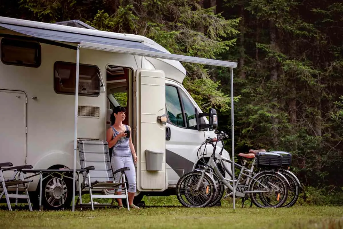 Una mujer está de pie en la puerta de su autocaravana blanca, aparcada en un sitio donde hay sillas y bicicletas expuestas fuera.