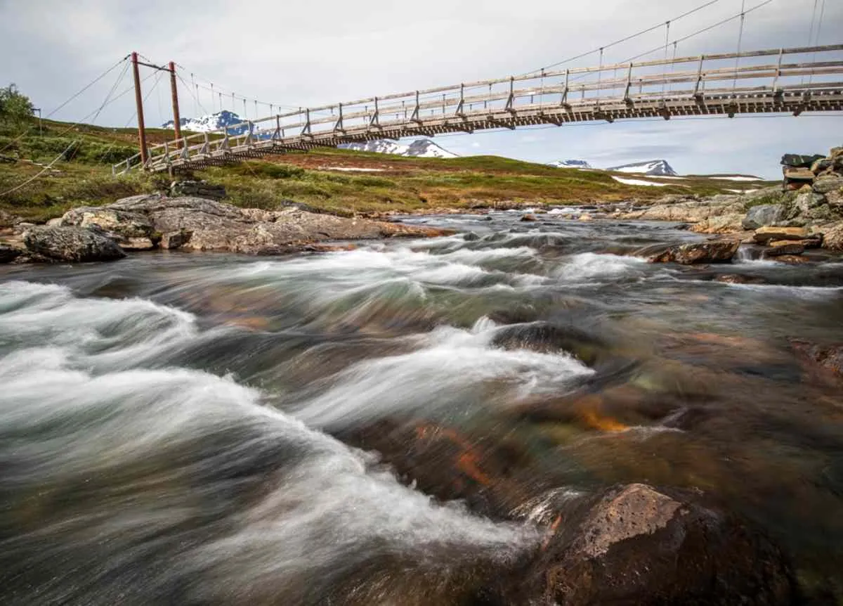 Vista d’insieme di un ponte di legno su un fiume con correnti molto forti, immerso in un paesaggio naturale con montagne innevate sullo sfondo.