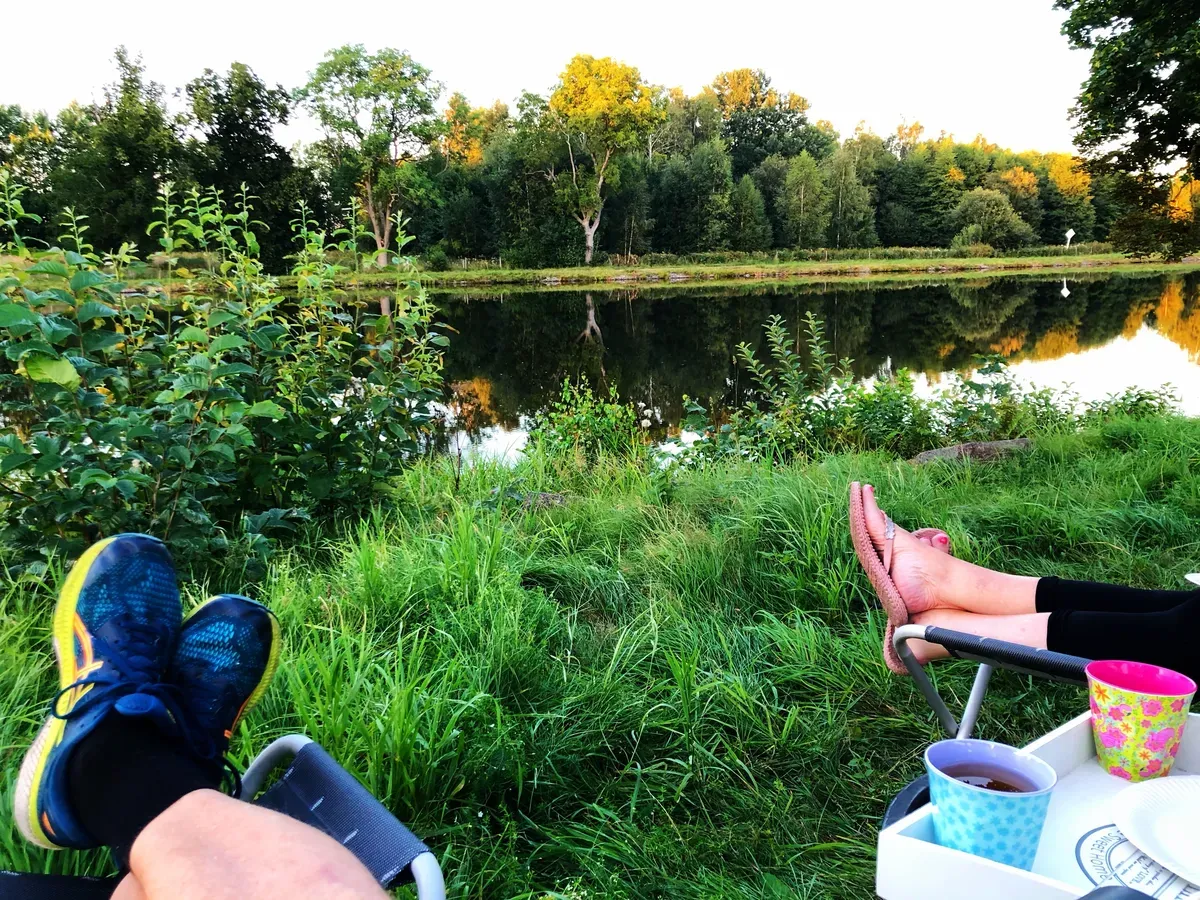 Dos personas relajándose en sillas de camping con los pies en alto y bebidas en una bandeja, contemplando las tranquilas aguas del canal de Göta a la hora dorada.