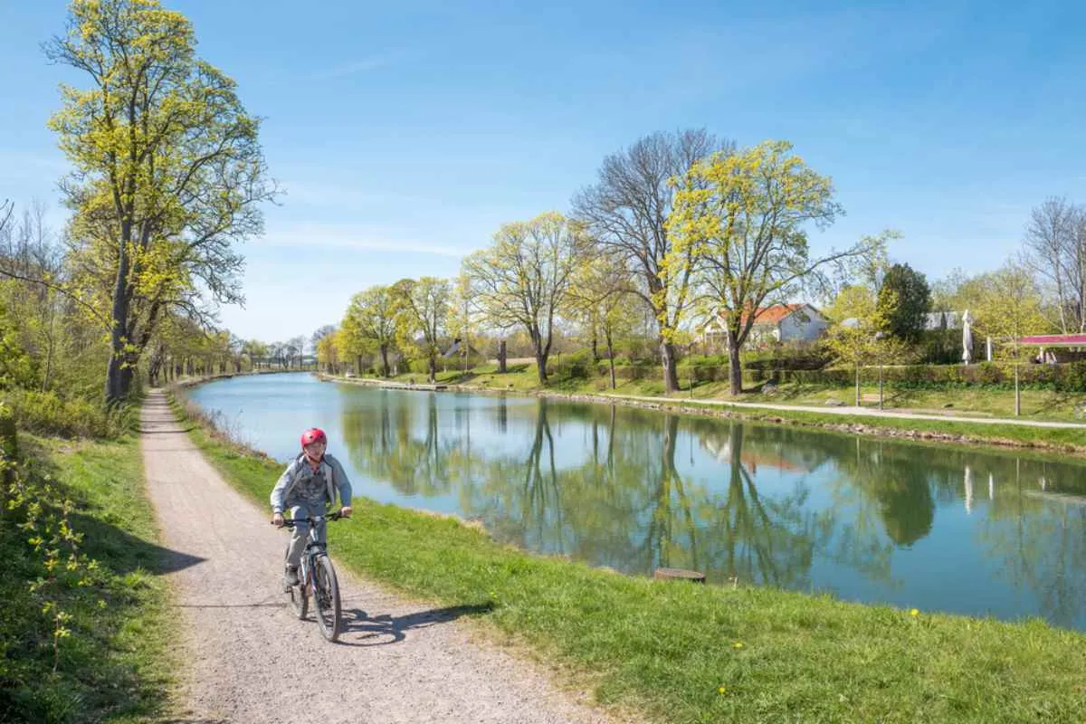 Una persona en bici por el camino de sirga del canal de Göta en un día soleado, con el agua tranquila reflejando los árboles verdes y el cielo azul.