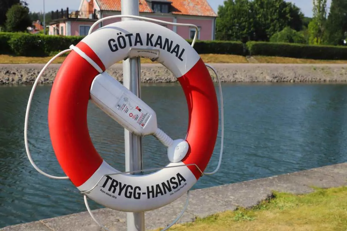 Primer plano de un salvavidas rojo y blanco con las inscripciones «Göta Kanal» y «Trygg-Hansa» junto al canal en un soleado día de verano.