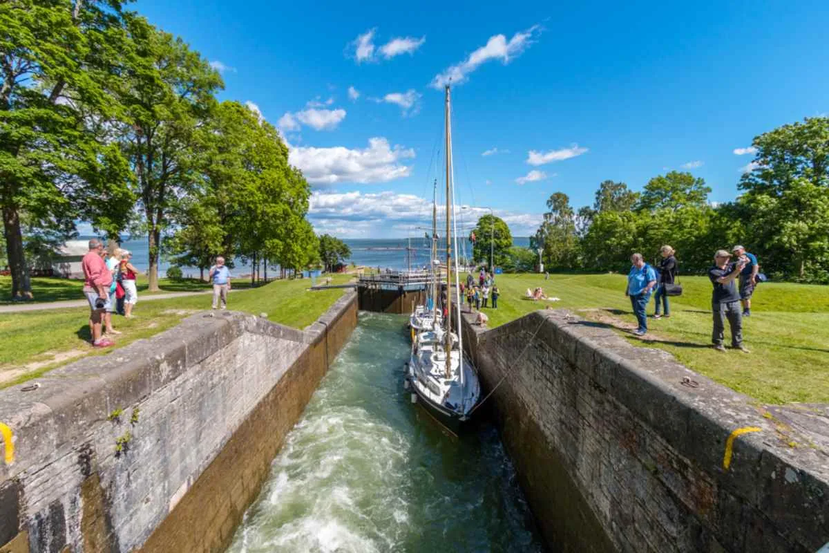 Un velero atraviesa una esclusa del canal de Göta mientras los visitantes lo observan desde las orillas cubiertas de hierba y los muros de piedra en un día soleado.