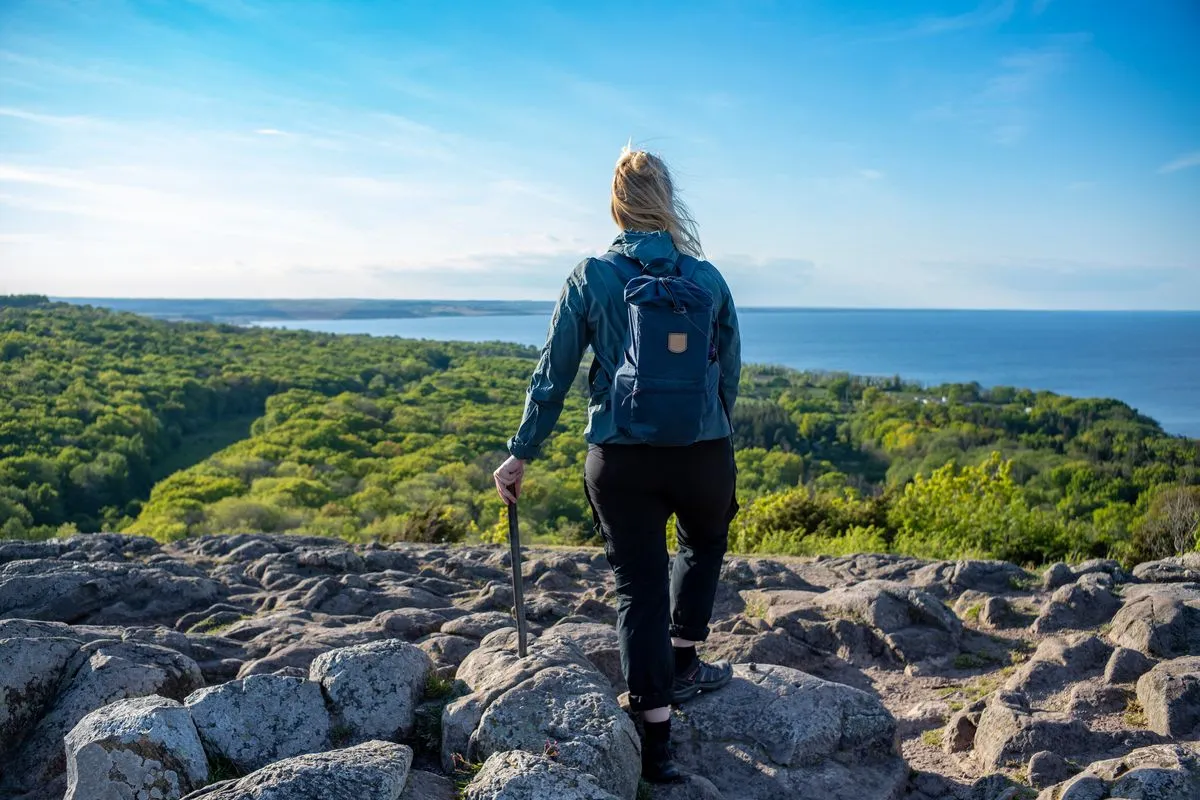 Una donna in abbigliamento da trekking e con l’attrezzatura da escursione è in piedi di spalle alla fotocamera e guarda intensamente l’orizzonte sopra alcune rocce.