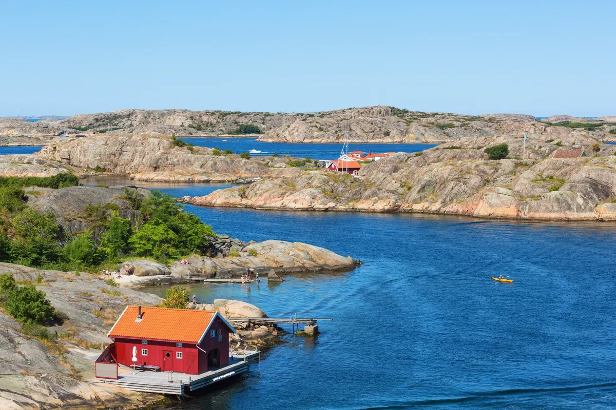 Una rimessa per barche rossa sulla costa rocciosa dell’arcipelago svedese, con acque blu e tranquille e casette sparse qua e là.