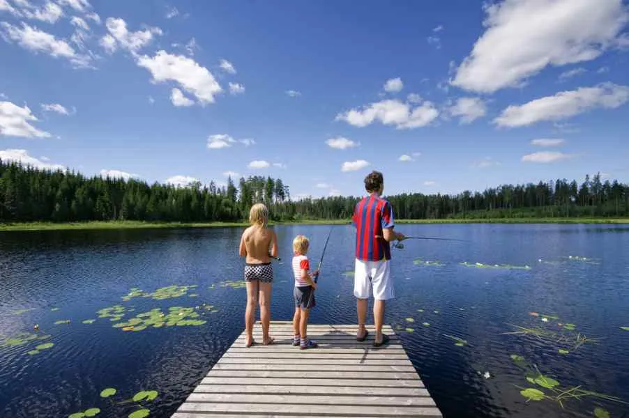 Una famiglia composta da un papà con una camicia blu e bianca e dai suoi bambini piccoli, che non la indossano, pesca su un molo di legno davanti a un lago svedese.