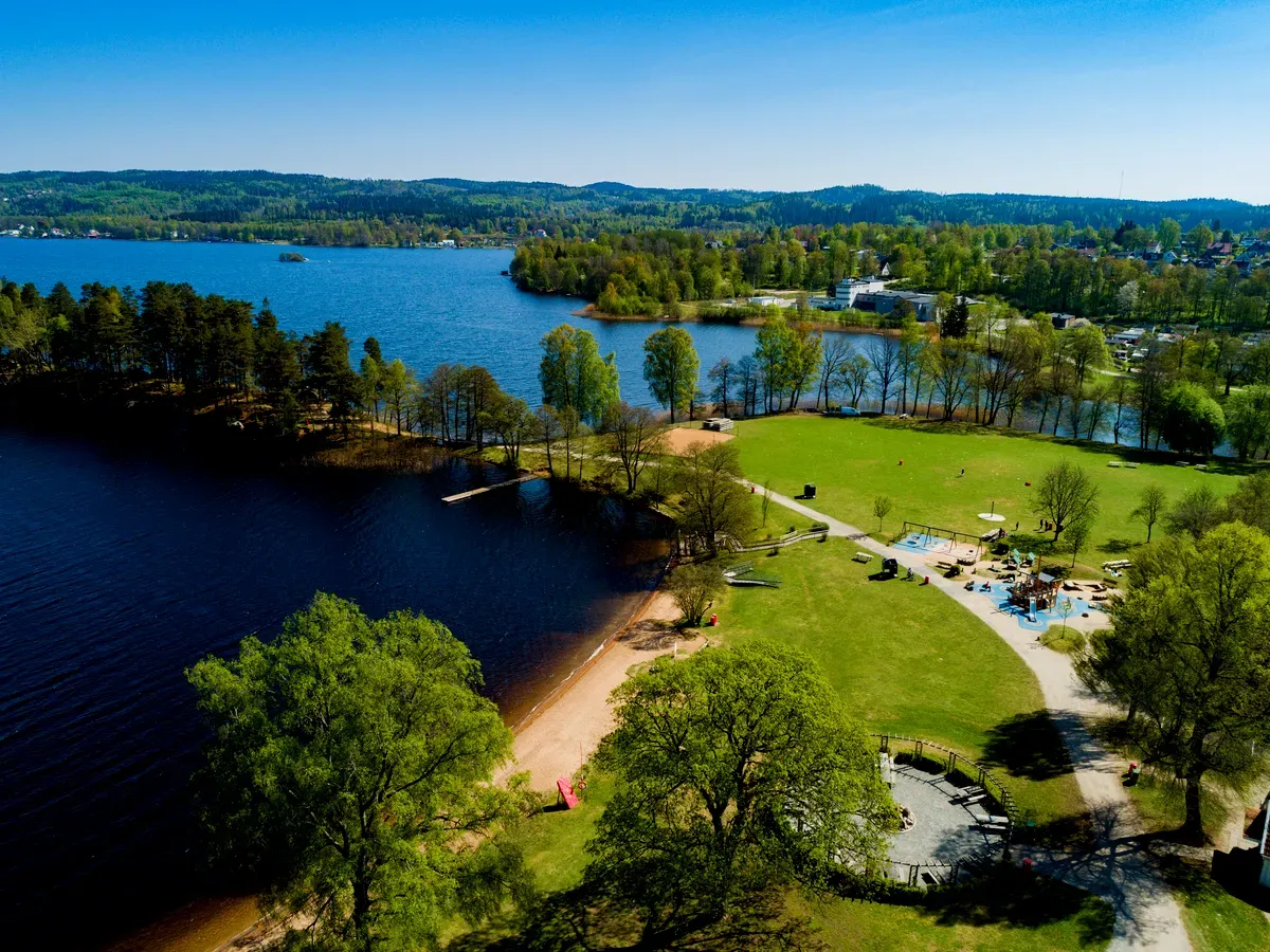 Vue aérienne d'un parc au bord d'un lac à Borås, avec une plage de sable, une jetée, des aires de jeux et des sentiers de promenade, par une journée ensoleillée.