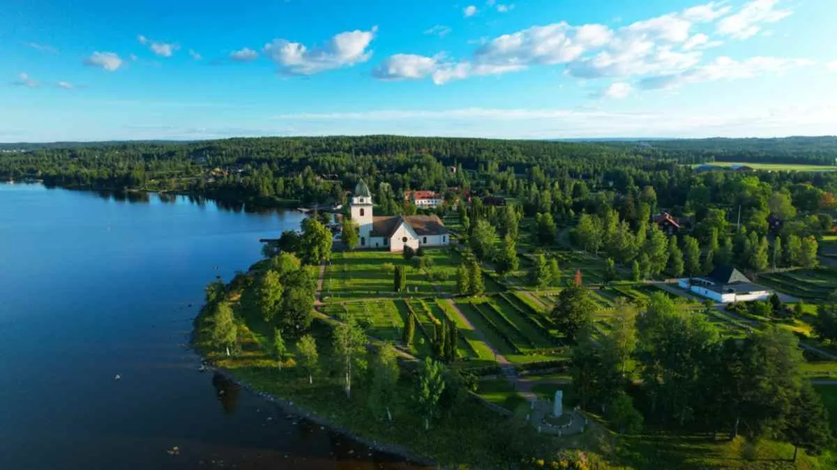 Vue aérienne d'une église de campagne blanche et de jardins à motifs au bord d'un lac paisible, entourés par la forêt en Dalécarlie, en Suède.