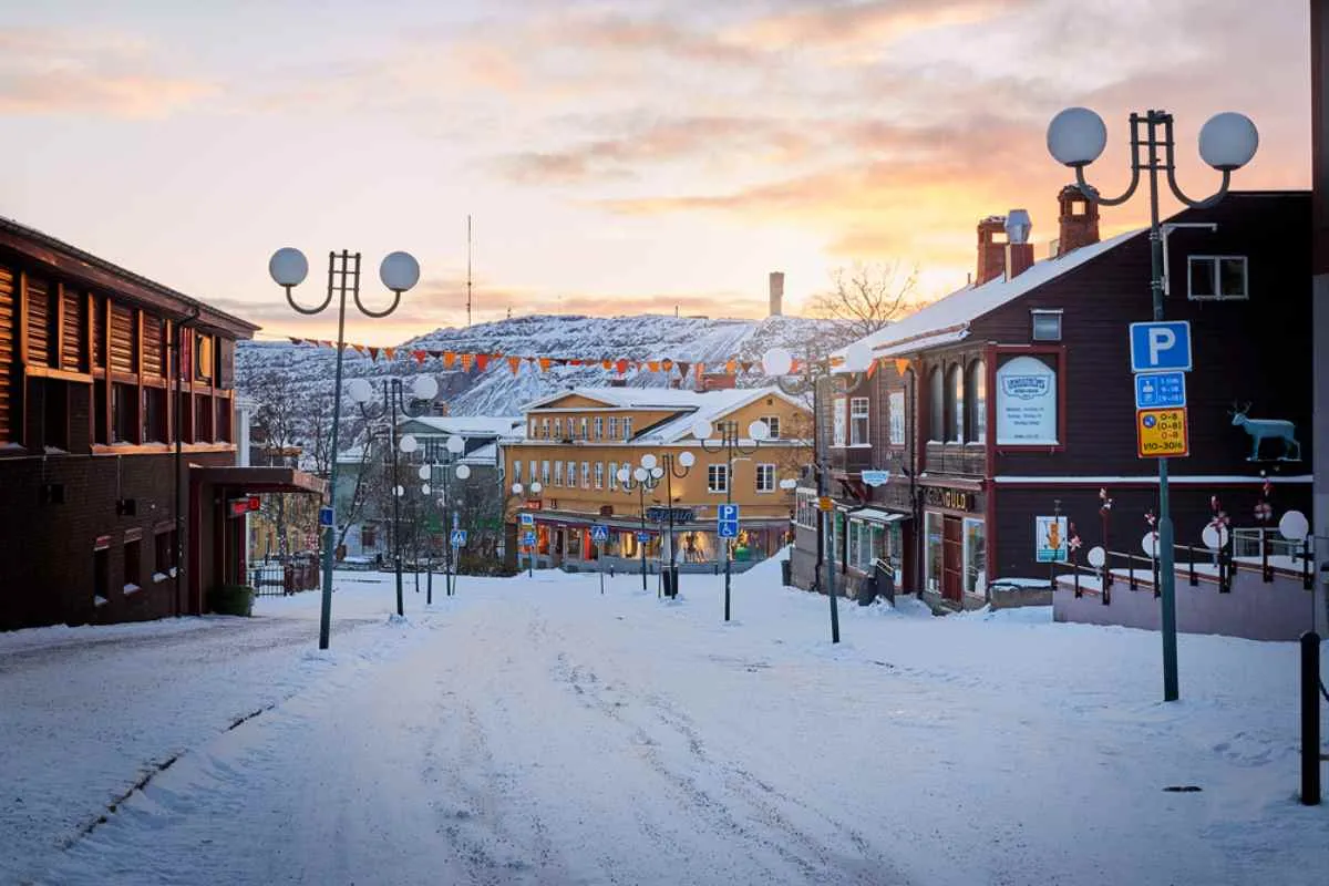 Rue principale enneigée de Kiruna au coucher du soleil, bordée de bâtiments en bois et de lampadaires, avec des collines en arrière-plan.