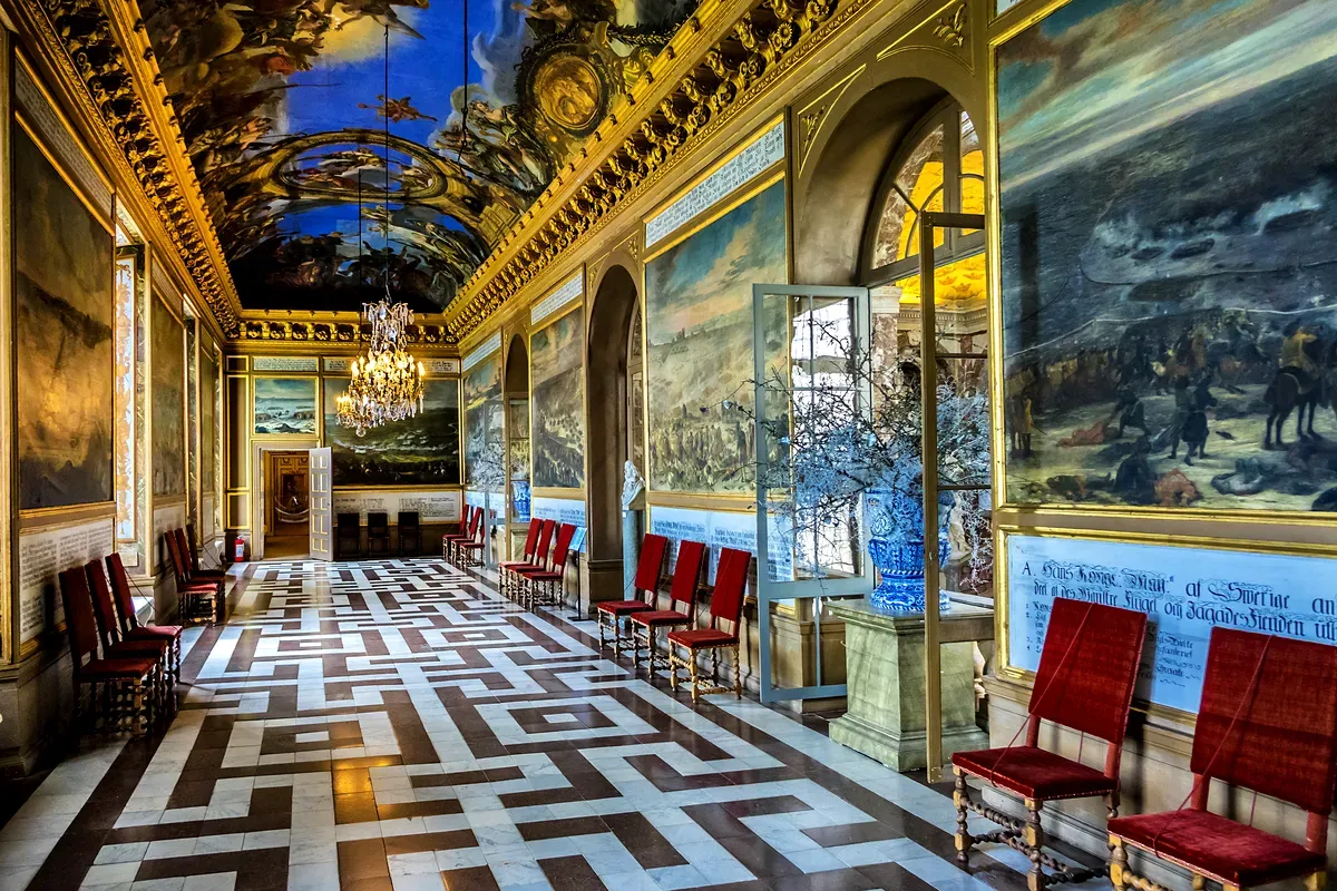 Interior del Palacio de Drottningholm, donde destaca un gran salón con pinturas ornamentadas en el techo, lámparas de araña y lujosas sillas rojas