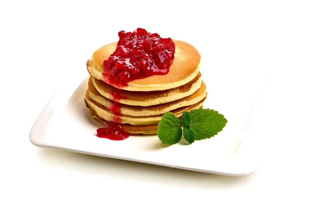 Une pile de pancakes moelleux nappés de sauce aux airelles rouges, agrémentés d’une feuille de menthe fraîche sur une assiette carrée blanche.
