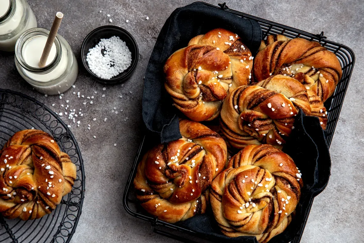 Une corbeille de brioches à la cannelle suédoises tout juste sorties du four, saupoudrées de sucre perlé, accompagnées de bouteilles de lait et d’un bol de sucre posés sur un plan de travail foncé