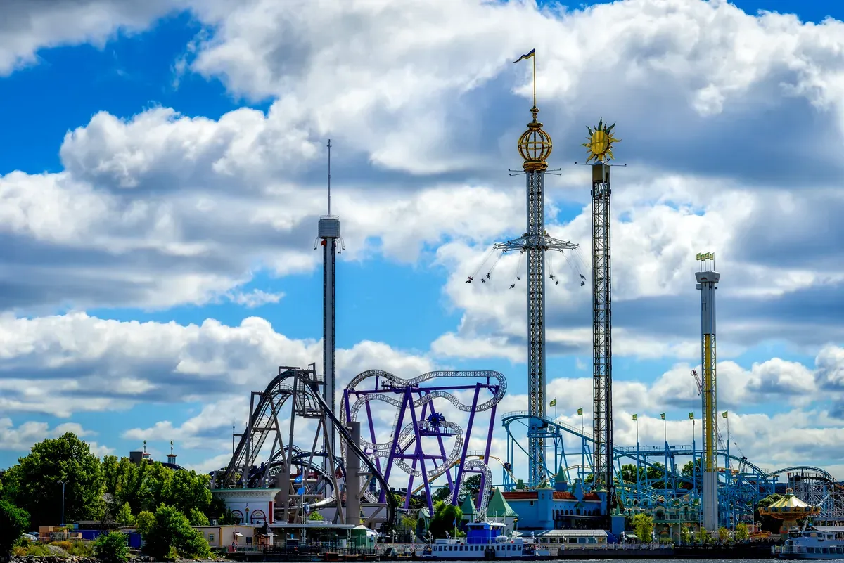 Una imagen llena de vida del parque de atracciones Gröna Lund en Estocolmo, con varias montañas rusas y atracciones de gran altura con un cielo azul y nubes blancas y esponjosas de fondo.