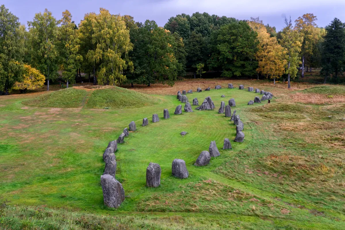 Una vista aerea del tumulo funerario di Anundshög e della formazione a forma di nave in pietra a Västerås, in Svezia, circondati da una vegetazione rigogliosa e da alberi con le foglie autunnali.