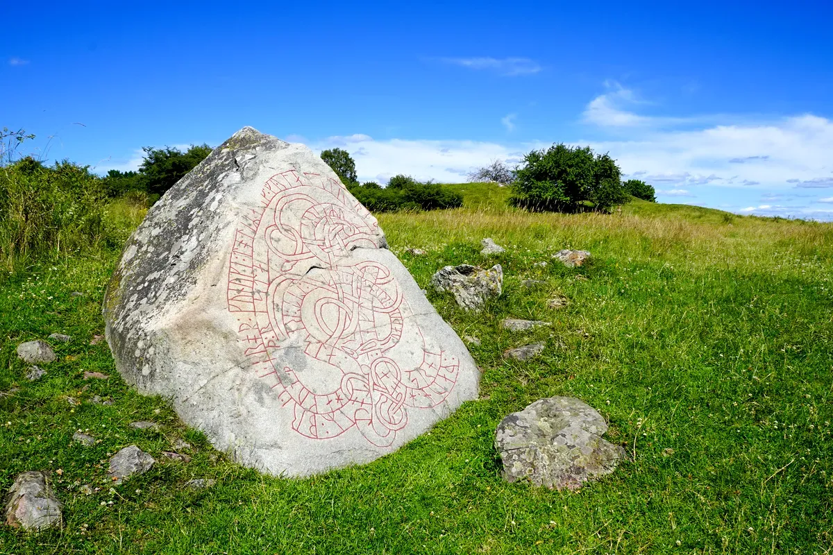 Una piedra rúnica vikinga en Birka, Suecia, erigida en un prado bajo un cielo azul claro, con intrincados grabados e inscripciones nórdicas visibles en su superficie.