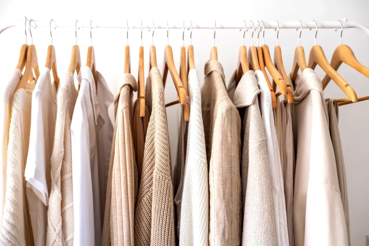 Una selezione di capi d’abbigliamento dai toni neutri, tra cui beige, bianco e crema, appesi con cura su grucce di legno appese a una rastrelliera di metallo bianco. I capi hanno texture diverse: alcuni sono in maglia, lino e altri tessuti morbidi, creando un’estetica coerente e minimalista. La palette cromatica complessiva è calda e rilassante, e riflette uno stile semplice e sobrio. La scena è ben illuminata, mettendo in risalto le trame e i colori dei capi, disposti in modo da creare un’esposizione equilibrata e armoniosa.