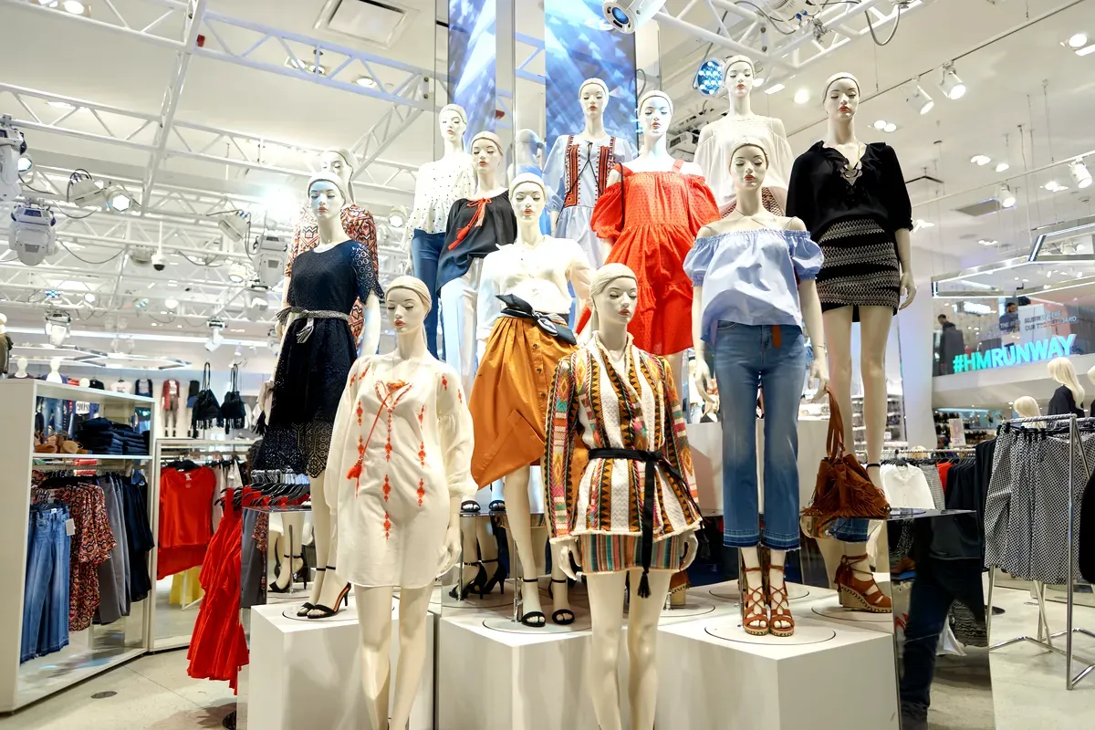 Una vetrina con manichini vestiti con vari outfit alla moda in un negozio H&M. I manichini sono disposti su più livelli e mettono in mostra una vasta gamma di stili di abbigliamento, tra cui abiti, gonne, jeans e camicette dai colori vivaci e con fantasie accattivanti. L’illuminazione brillante del negozio mette in risalto la moda trendy e contemporanea, con in sfondo l’insegna del negozio e altri espositori di abbigliamento. L’atmosfera generale è moderna ed energica, e riflette il fascino del marchio per un pubblico giovane e attento allo stile.