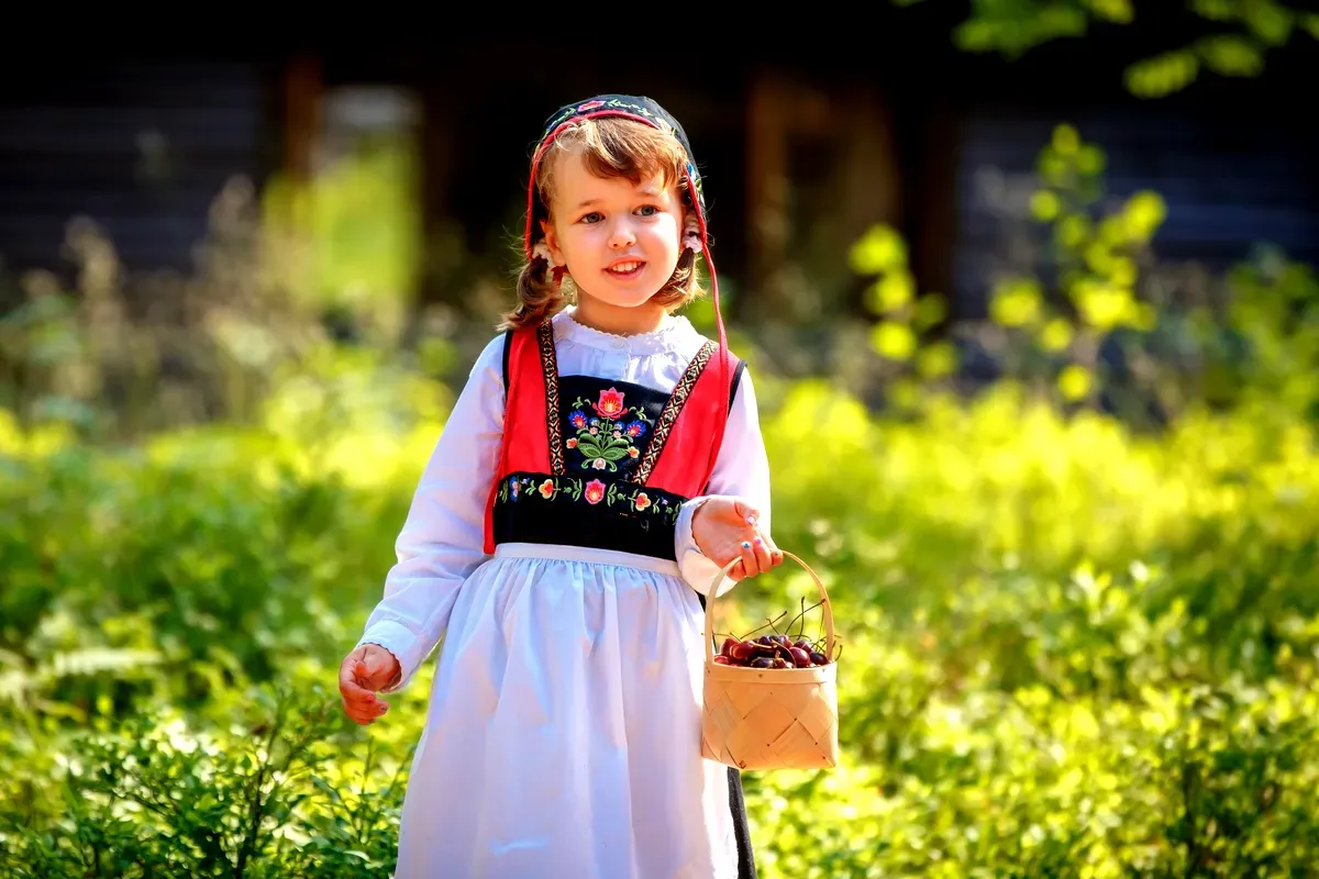Una ragazzina che indossa un abito tradizionale svedese è in piedi all’aperto, con in mano un piccolo cesto pieno di bacche. Indossa un abito colorato con intricati ricami floreali sul corpetto e un foulard che si abbina al vestito. La luce del sole filtra tra gli alberi, illuminandola con un bagliore caldo e creando una scena pittoresca e affascinante del patrimonio culturale svedese. Lo sfondo è caratterizzato da una vegetazione rigogliosa, che esalta l’atmosfera tranquilla e idilliaca.