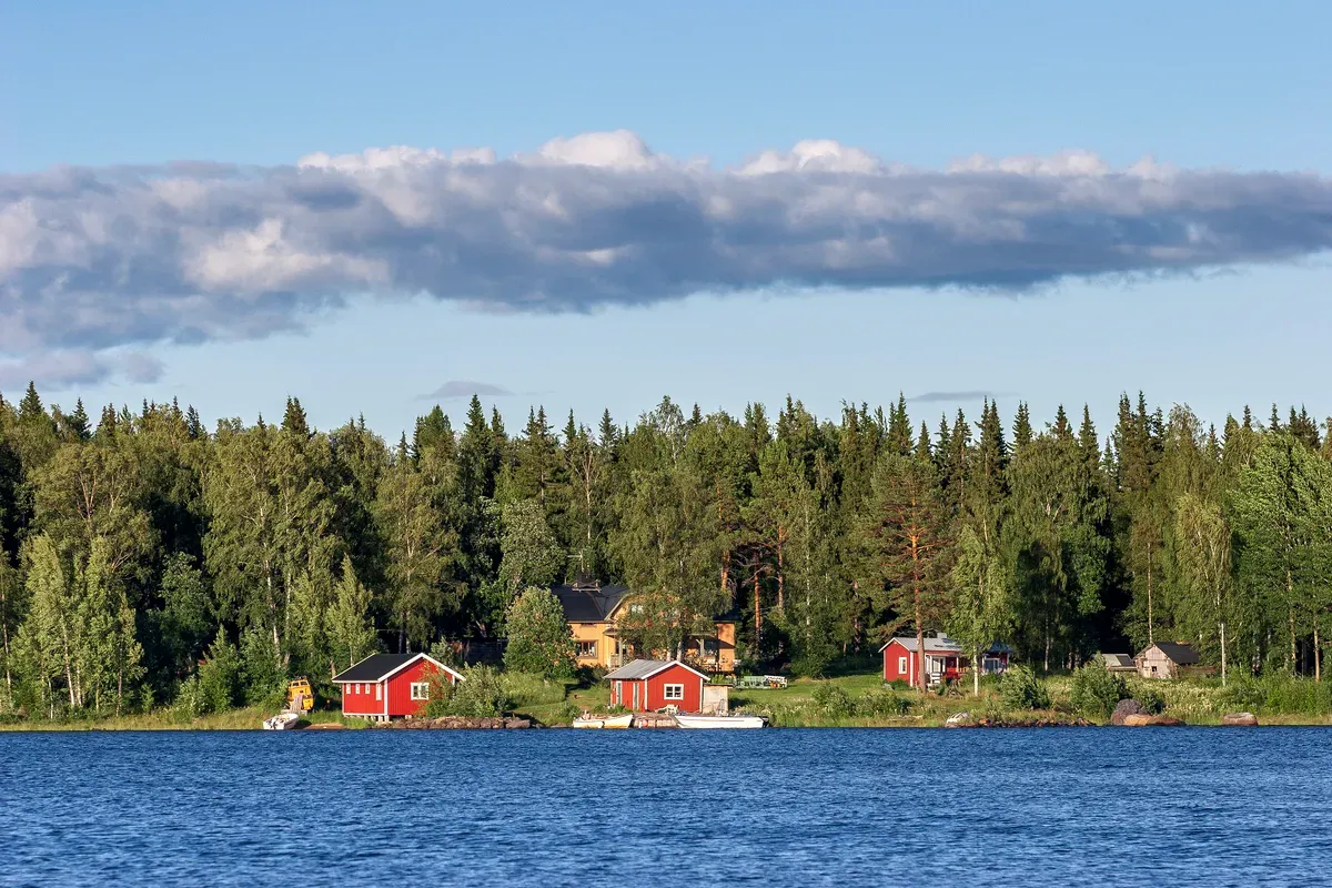 Vista panorámica de las tradicionales casitas rojas de madera suecas, enclavadas entre densos bosques verdes a orillas de un tranquilo lago azul. Las casitas están rodeadas de frondosos árboles y hay unas cuantas barcas pequeñas amarradas cerca de la orilla. El cielo está despejado, con algunas nubes dispersas, lo que crea un ambiente tranquilo e idílico, típico de la campiña sueca. La escena transmite una sensación de tranquilidad y conexión con la naturaleza, característica de la Suecia rural.
