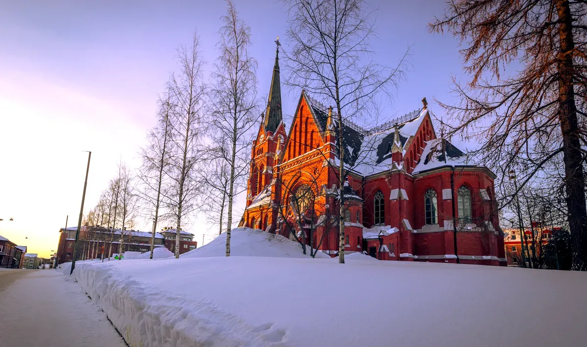 La catedral de Luleå en Suecia durante el invierno: una iglesia histórica de ladrillo rojo iluminada por la puesta de sol, con los alrededores cubiertos de nieve.