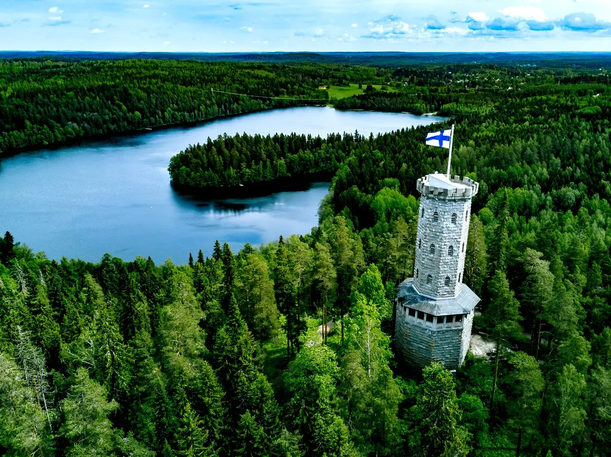 Vista aérea de la histórica torre de observación de piedra con la bandera finlandesa, rodeada de densos bosques verdes y un tranquilo lago azul en Finlandia.
