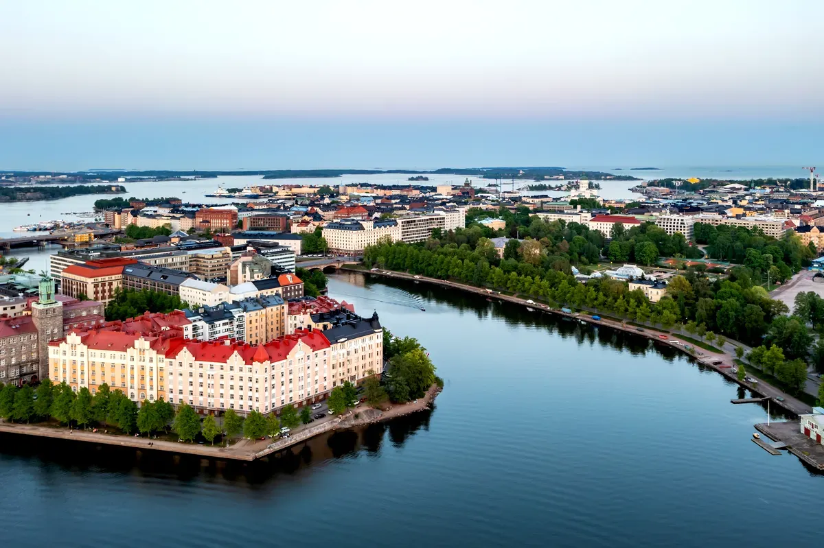 Una vista aérea de Helsinki, Finlandia, que muestra la mezcla única de arquitectura histórica y moderna de la ciudad. La imagen capta el vibrante paisaje urbano con edificios de colores, sobre todo las construcciones de tejados rojos en primer plano, rodeadas de frondosos árboles y cursos de agua. La ciudad se extiende hacia el horizonte, con el mar Báltico y pequeñas islas visibles a lo lejos, creando una pintoresca escena costera bajo un cielo azul claro y suave.