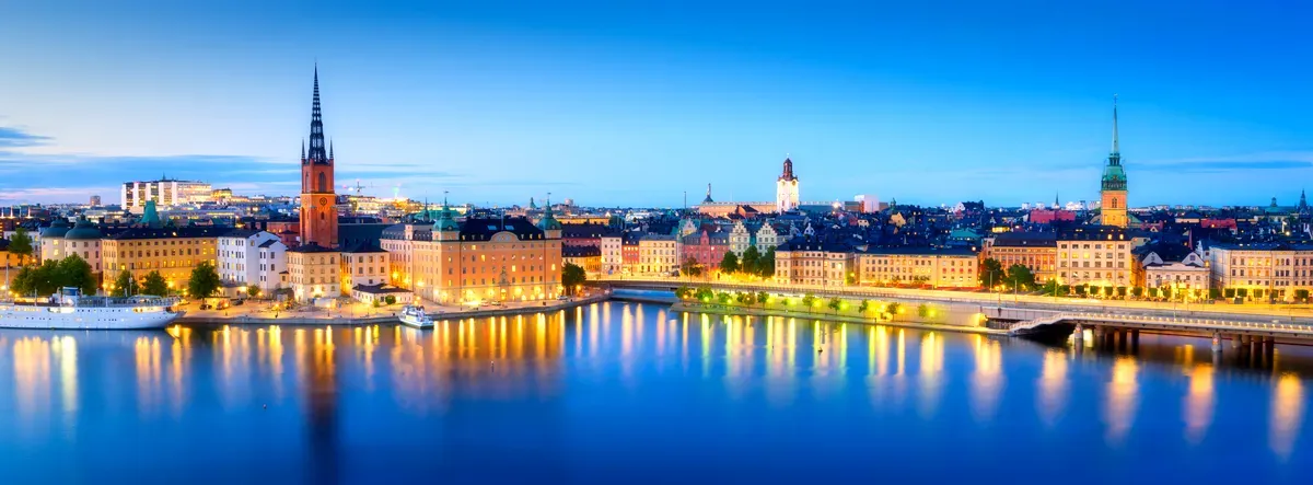 Vista panorámica del histórico perfil urbano de Estocolmo al atardecer, con sus emblemáticas agujas, edificios iluminados y reflejos en las tranquilas aguas del mar Báltico.